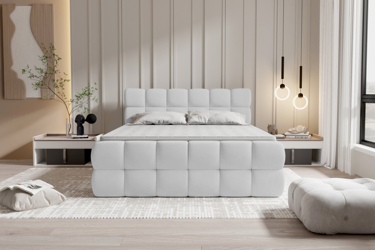 Modernes, gepolstertes Bett in Weiß mit quadratisch gestepptem Kopf- und Fußteil in einem hellen Schlafzimmer, frontal aus Augenhöhe fotografiert; rechts hängende Ring-Pendelleuchten und links Nachttisch mit Deko.