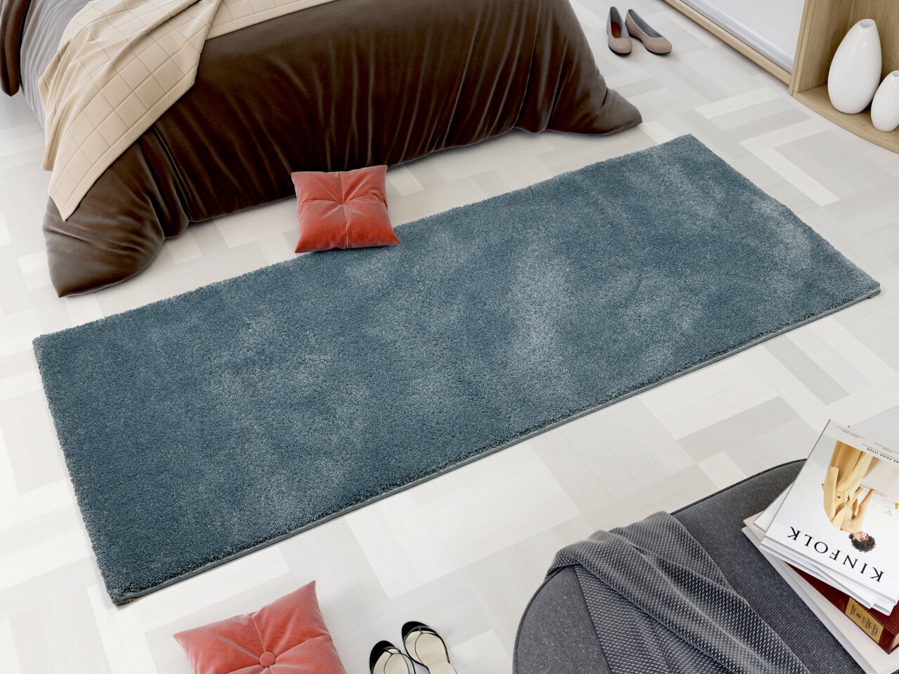 Rechteckiger, blauer Uni-Teppich in einem modernen Schlafzimmer, von oben betrachtet. Der Teppich liegt auf einem hellen Holzboden neben einem Bett mit brauner Decke und roten Kissen.