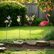 Frontalansicht auf dekorative Gartenstecker im Rasen: bunte Schmetterlings- und Blumenmotive sowie ein pinker Flamingo vor Hecke und Holzzaun im Sonnenlicht.