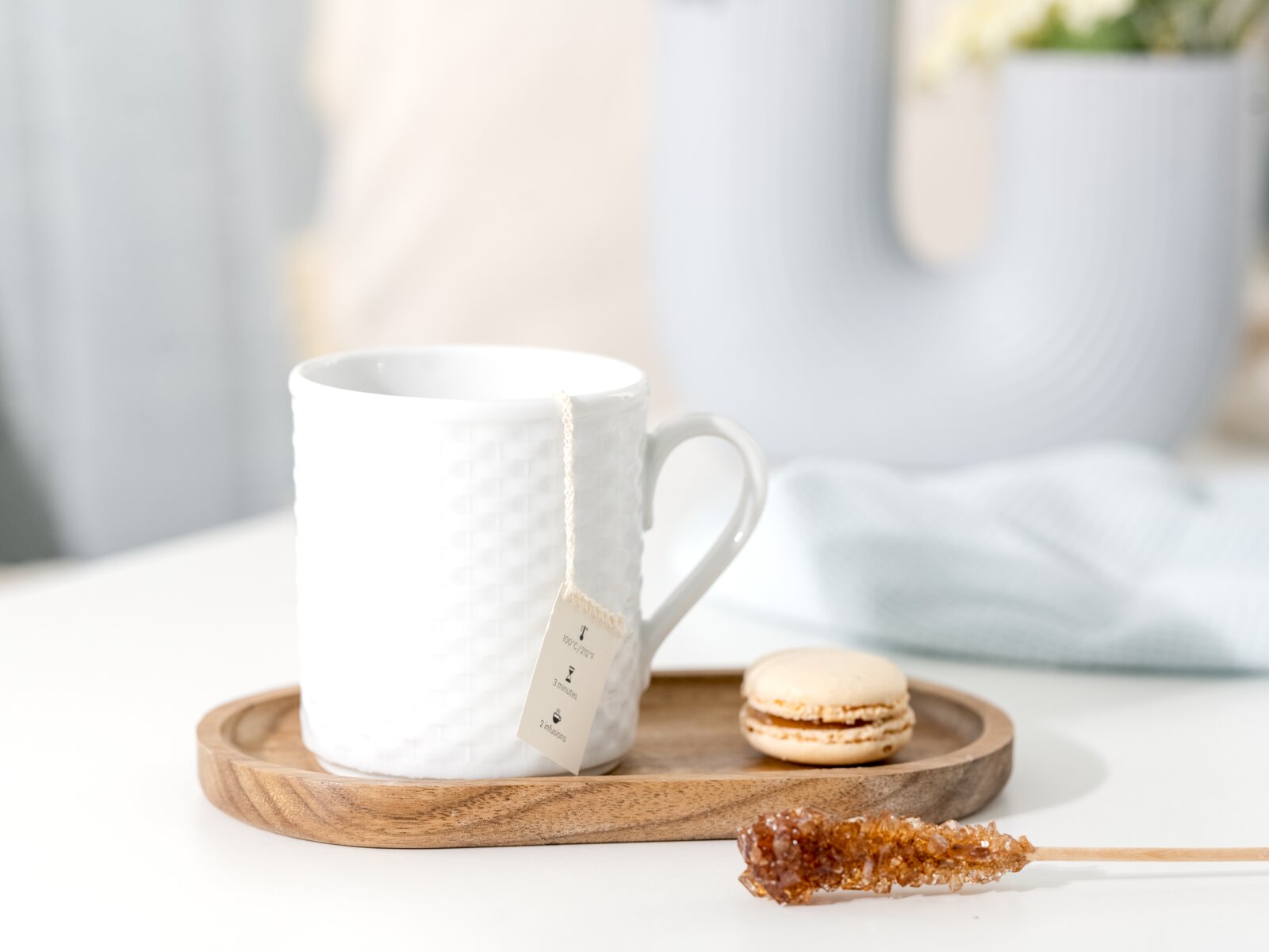 Weiße Keramiktasse mit Teebeutel auf Holztablett, daneben Macaron und Kandiszuckerstick; Perspektive: frontale Nahaufnahme auf Tischhöhe mit leichtem Seitenwinkel