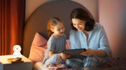 Eine Mutter und ihre Tochter sitzen auf einem Bett und schauen gemeinsam auf ein Tablet. Auf dem Nachttisch neben ihnen steht eine leuchtende Akku-Nachtleuchte in Form eines Welpen. Die Szene ist aus einer seitlichen Perspektive aufgenommen.