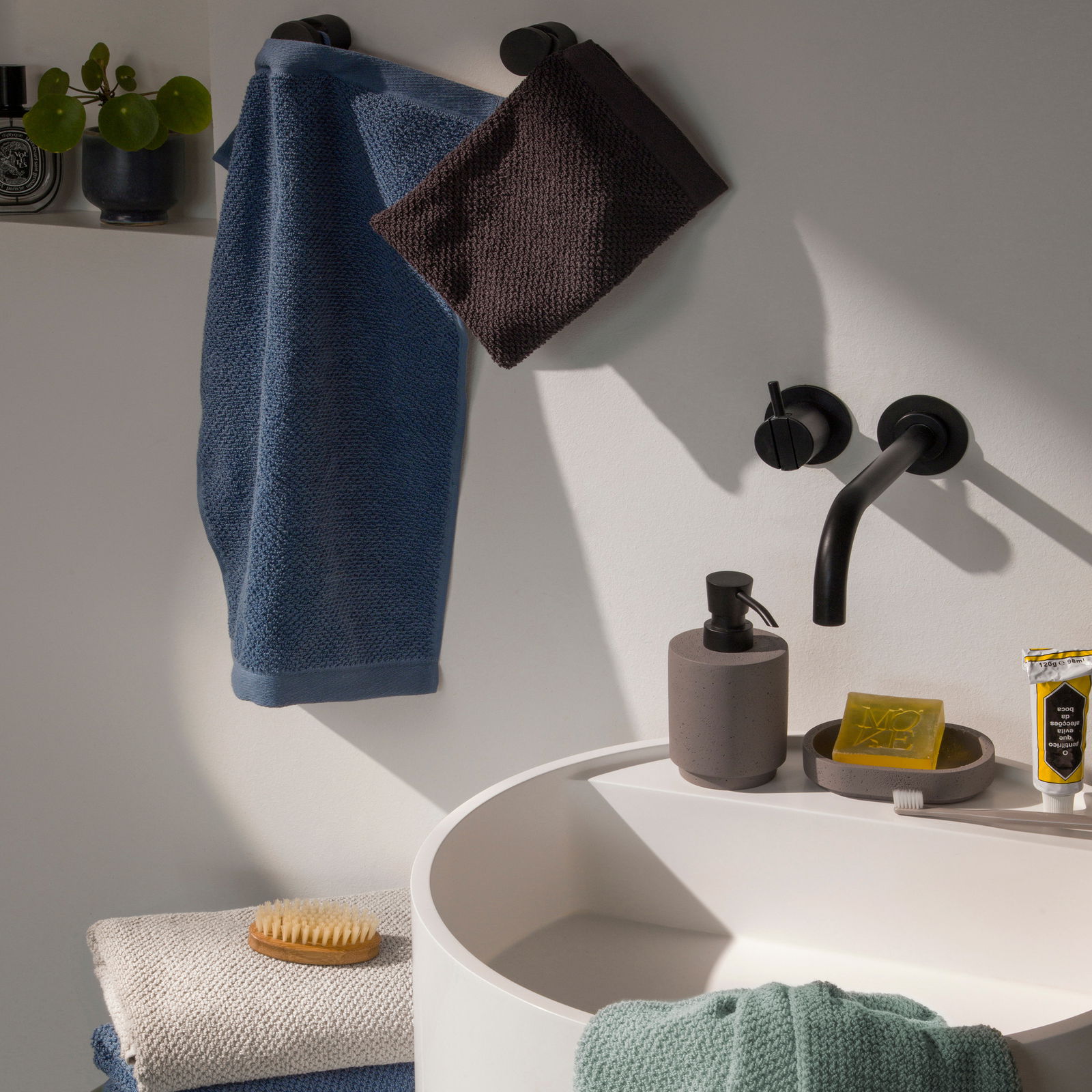 MÖVE Handdoekenset, 10-delig BLISS Badezimmer mit Handtüchern in verschiedenen Farben, aufgehängt an schwarzen Haken, mit Waschbecken und Seifenspender, seitliche Perspektive.
