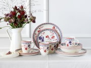 Blumenverziertes Porzellan-Geschirrset mit Tellern, Schalen und Tassen in Weiß mit roten und blauen Blütenmotiven, frontal in leichter Schrägansicht auf einem gedeckten Tisch neben einer weißen Vase mit Blumen.