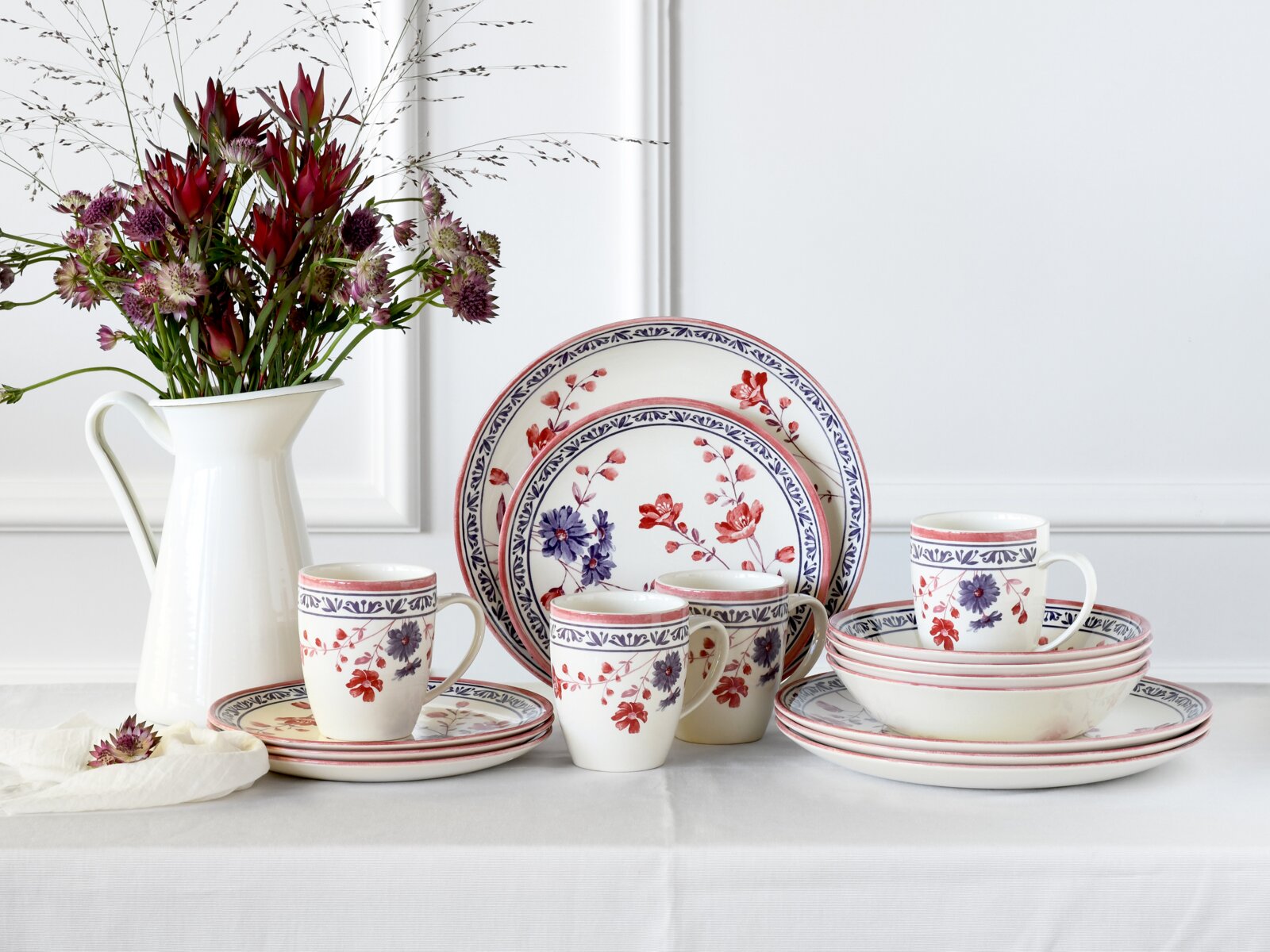 creaTable Kombiservice 16-tlg COTTAGE Blumenverziertes Porzellan-Geschirrset mit Tellern, Schalen und Tassen in Weiß mit roten und blauen Blütenmotiven, frontal in leichter Schrägansicht auf einem gedeckten Tisch neben einer weißen Vase mit Blumen.