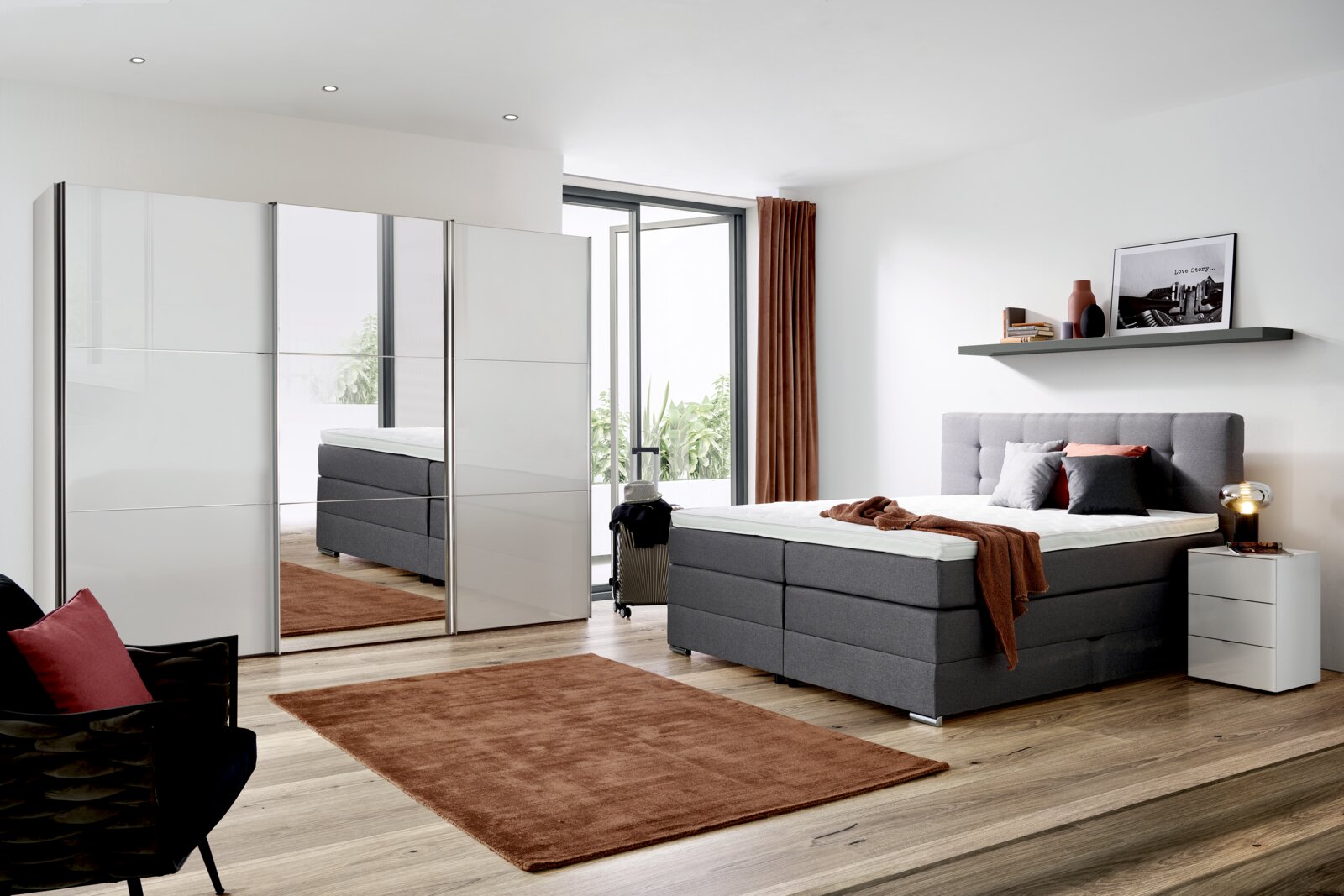 Modernes Schlafzimmer mit einem grauen Boxspringbett, das von der Seite gezeigt wird. Das Bett ist mit Kissen und einer Decke dekoriert. Im Hintergrund sind ein Kleiderschrank mit Spiegel, ein Nachttisch und ein Regal mit Dekorationen zu sehen.