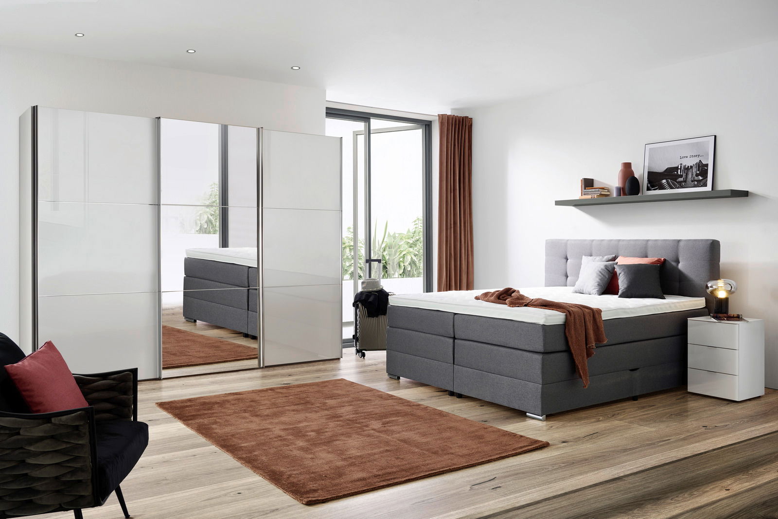 Modernes Schlafzimmer mit einem grauen Boxspringbett, das von der Seite gezeigt wird. Das Bett ist mit Kissen und einer Decke dekoriert. Im Hintergrund sind ein Kleiderschrank mit Spiegel, ein Nachttisch und ein Regal mit Dekorationen zu sehen.