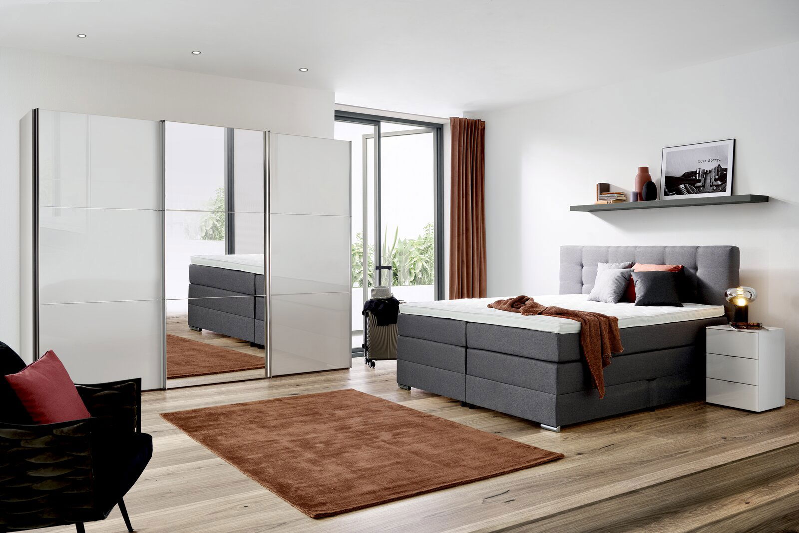Modernes Schlafzimmer mit einem grauen Boxspringbett, das von der Seite gezeigt wird. Das Bett ist mit Kissen und einer Decke dekoriert. Im Hintergrund sind ein Kleiderschrank mit Spiegel, ein Nachttisch und ein Regal mit Dekorationen zu sehen.