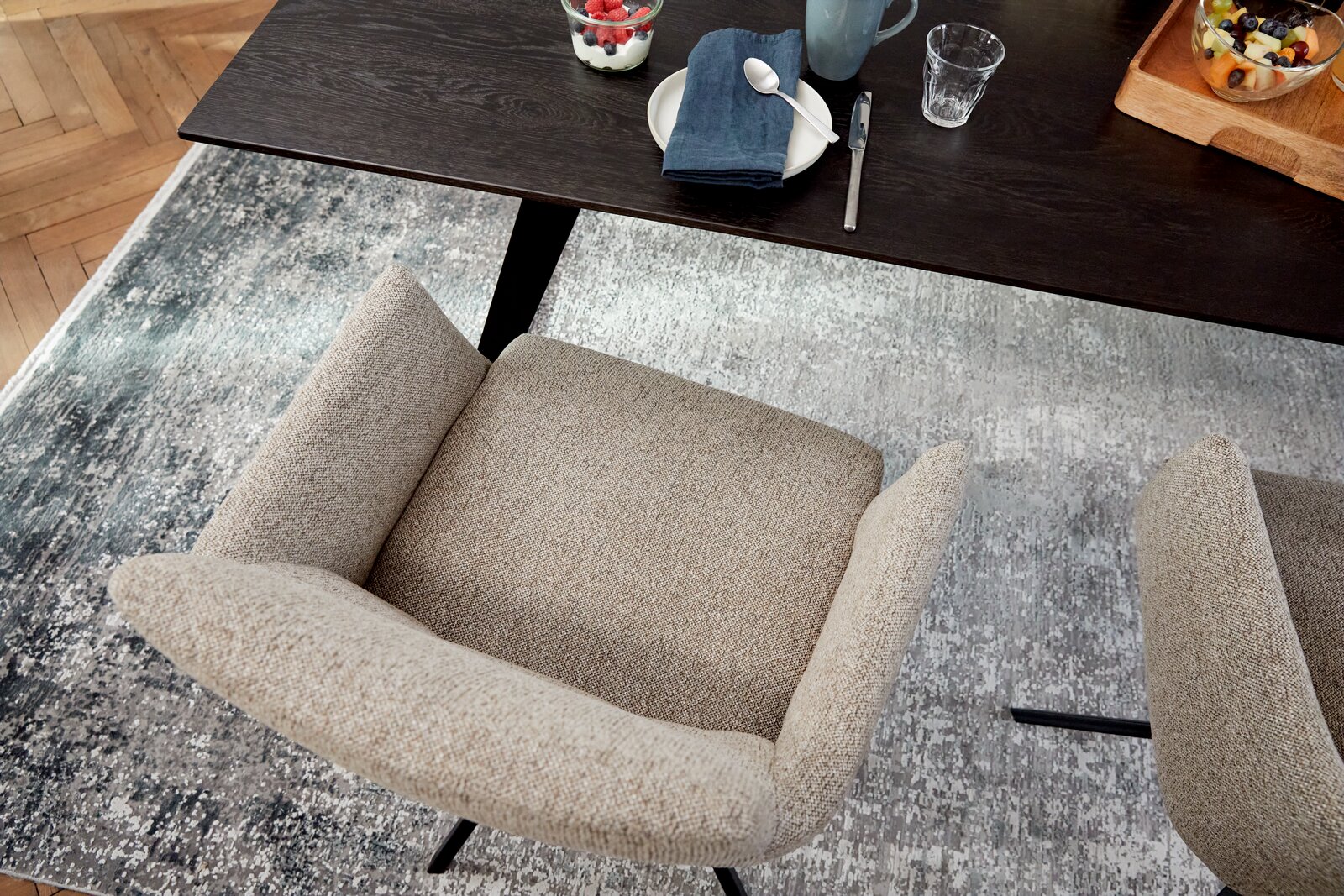 Vogelperspektive auf einen beigen Sessel mit Armlehnen, der an einem dunklen Holztisch steht, auf einem gemusterten Teppich.