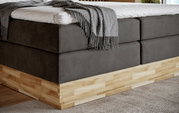 Close-up van een modern boxspringbed met grijze stoffen bekleding en houten onderstel, gezien vanaf de zijkant.
