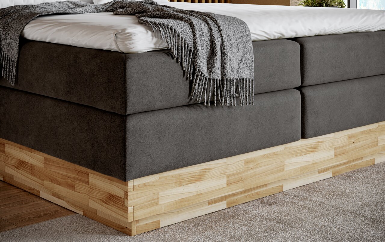 Close-up van een modern boxspringbed met grijze stoffen bekleding en houten onderstel, gezien vanaf de zijkant.