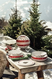 Ein festlich gedeckter Tisch im Freien mit einem dekorativen Geschirrset 'La Boule' in Rot und Weiß, umgeben von schneebedeckten Tannenbäumen. Die Perspektive zeigt das Geschirrset von der Seite.
