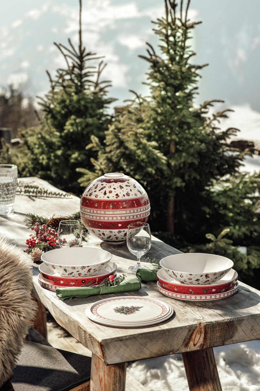 Ein festlich gedeckter Tisch im Freien mit einem dekorativen Geschirrset 'La Boule' in Rot und Weiß, umgeben von schneebedeckten Tannenbäumen. Die Perspektive zeigt das Geschirrset von der Seite.