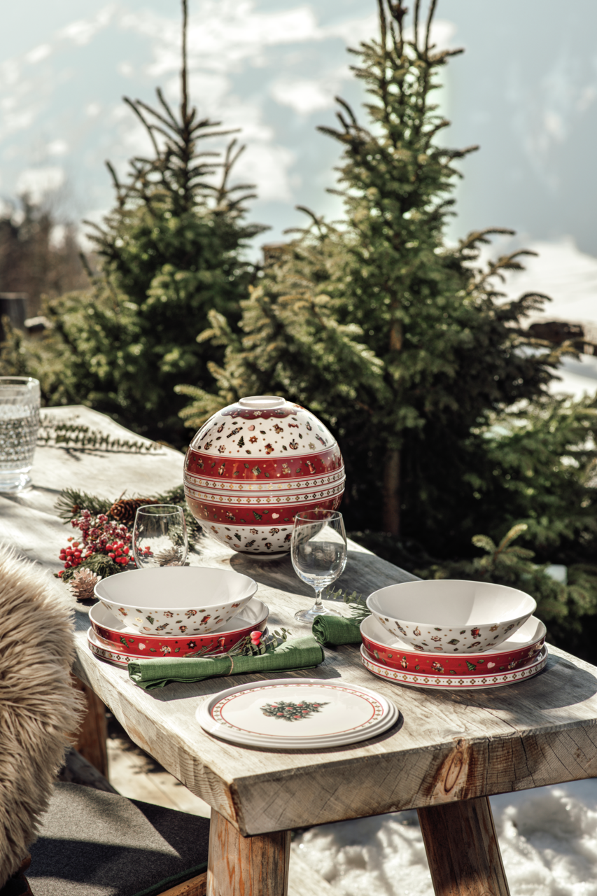 Ein festlich gedeckter Tisch im Freien mit einem dekorativen Geschirrset 'La Boule' in Rot und Weiß, umgeben von schneebedeckten Tannenbäumen. Die Perspektive zeigt das Geschirrset von der Seite.