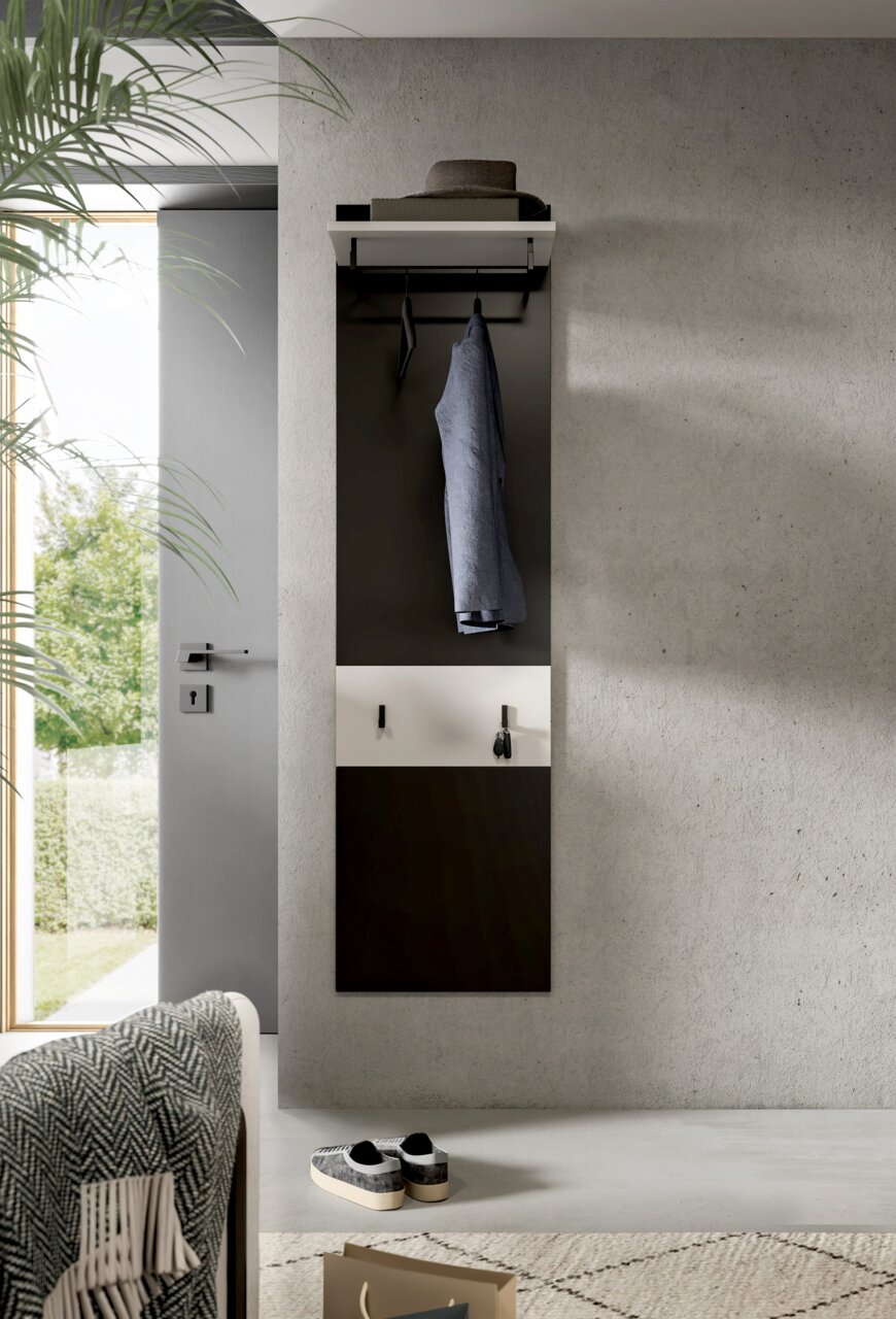 Modernes, wandmontiertes Garderobenpaneel in Schwarz und Weiß mit Ablage, Kleiderstange und Haken, frontal aus leicht seitlicher Perspektive im Flur an Betonwand inszeniert; Jacke hängt mittig, Hut liegt oben auf der Ablage.
