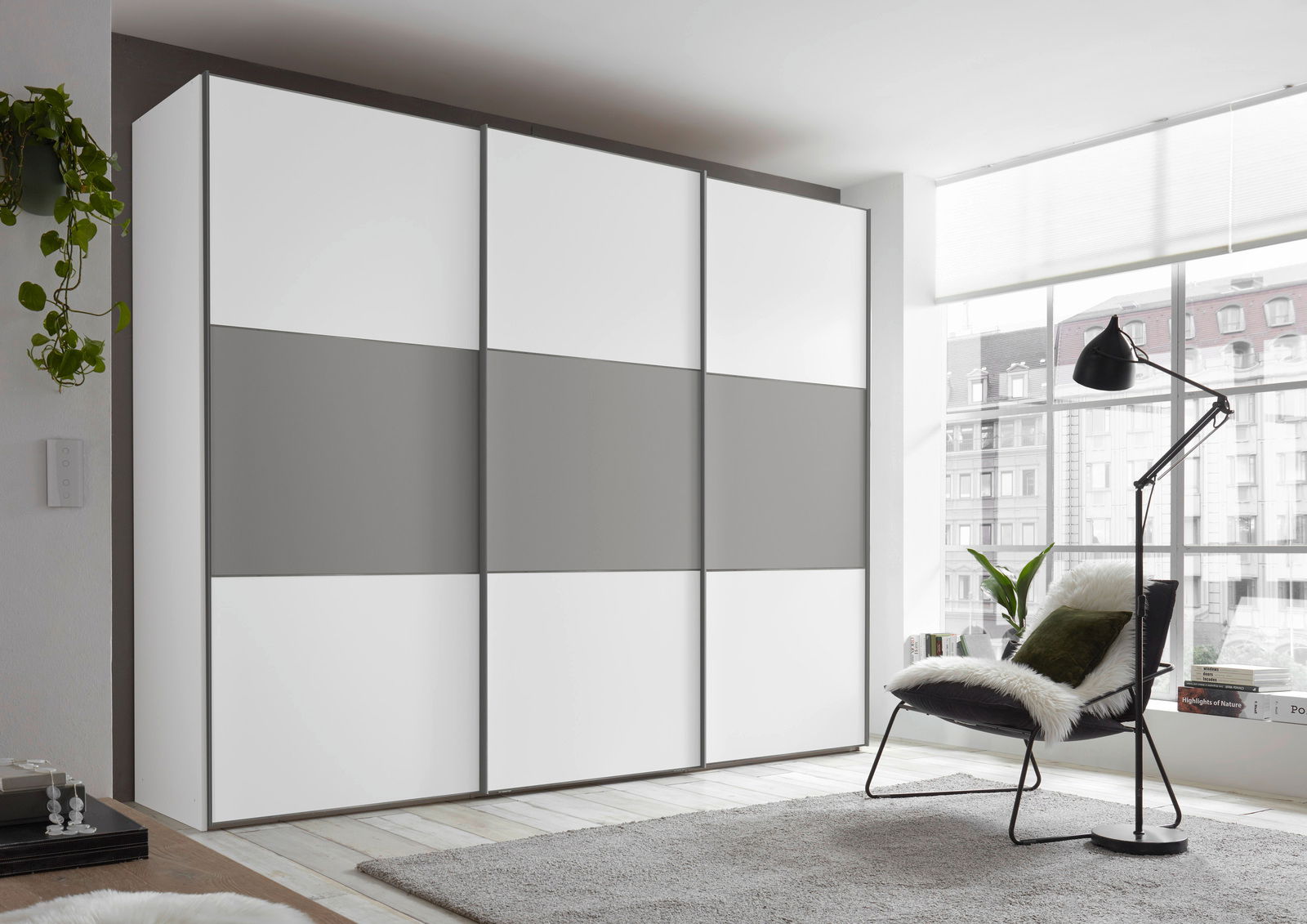 Moderner Schwebetürenschrank in Weiß und Grau, seitliche Perspektive, in einem hellen Raum mit großen Fenstern.