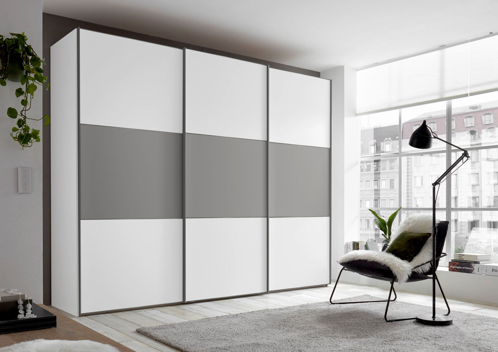 Moderano Schwebetürenschrank PRIMO Moderner Schwebetürenschrank in Weiß und Grau, seitliche Perspektive, in einem hellen Raum mit großen Fenstern.