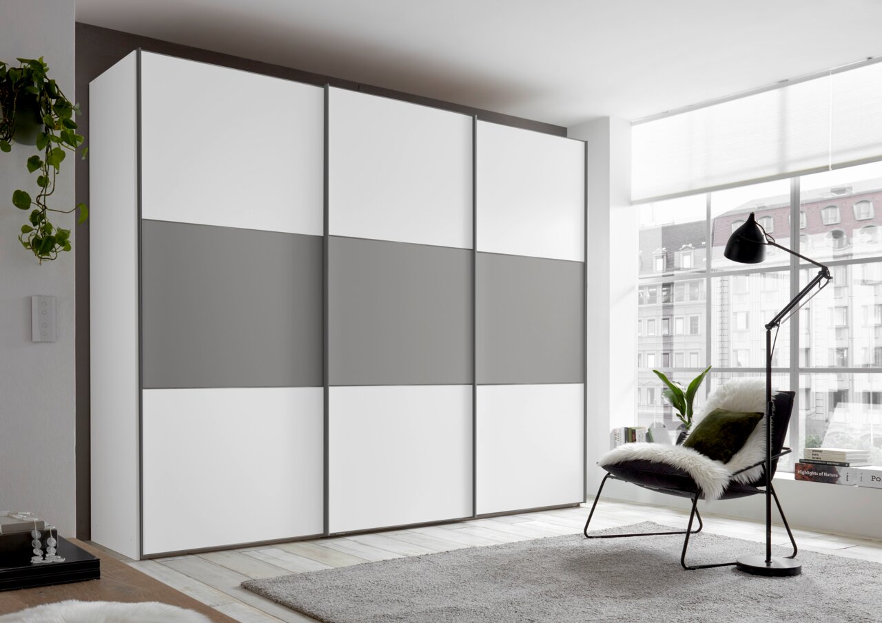 Moderner Schwebetürenschrank in Weiß und Grau, seitliche Perspektive, in einem hellen Raum mit großen Fenstern.