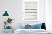 Doppelrollo mit Blende in einem modernen Schlafzimmer, frontal von innen fotografiert, mit einem Bett und Dekorationen im Vordergrund.