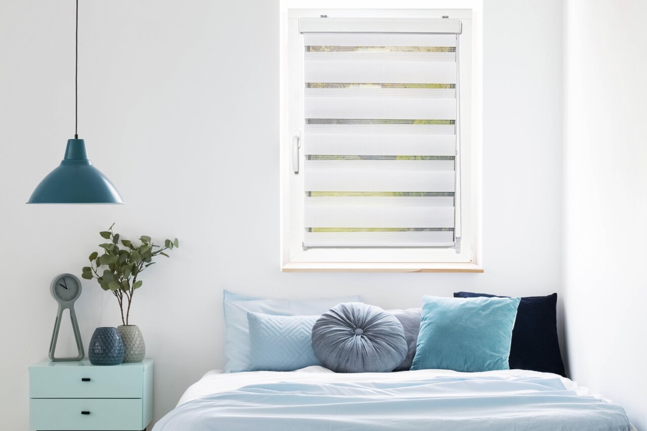 Een dubbelrolgordijn met een paneel in een moderne slaapkamer, van voren gefotografeerd vanuit de kamer, met een bed en decoraties op de voorgrond.