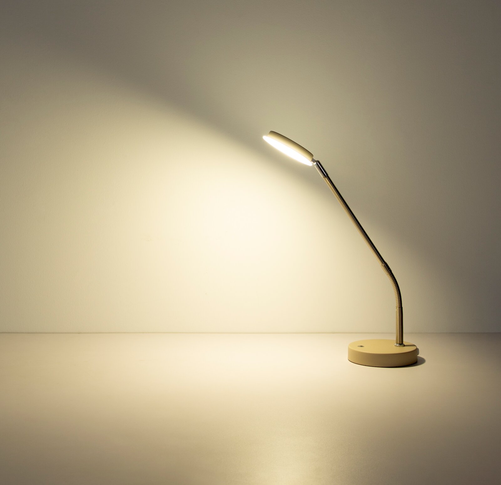 Moderne LED-Schreibtischlampe in Weiß mit rundem Standfuß und flexiblem Metallarm, eingeschaltet und nach links gerichtet, Seitenansicht auf einem hellen Tisch vor neutraler Wand