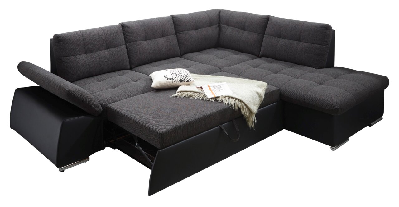Hoekbank met donkergrijze stoffen bekleding, uitgeschoven tot een bed, met kussens en deken, gezien vanuit een schuine hoek aan de voorkant.