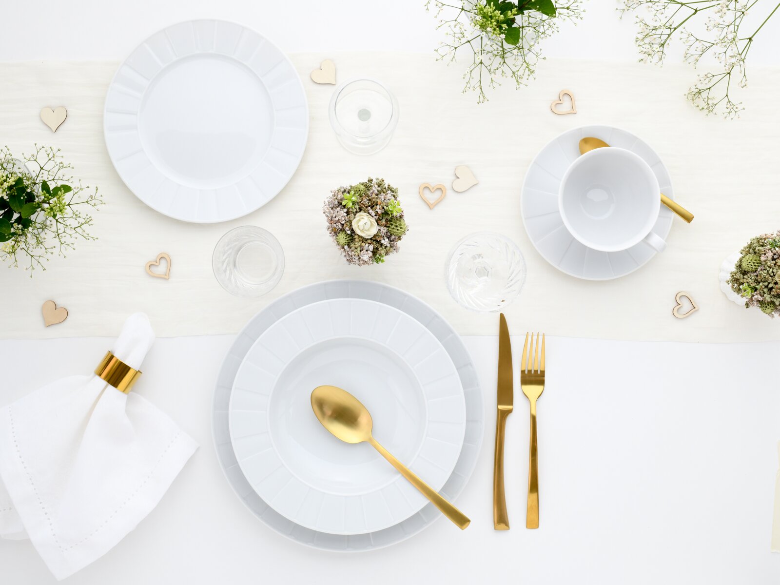Elegantes Tischgedeck in Weiß und Gold mit Porzellantellern, Tasse, goldfarbenem Besteck, Gläsern, Blumen und Herz-Deko auf hellem Tischläufer, Draufsicht von oben.