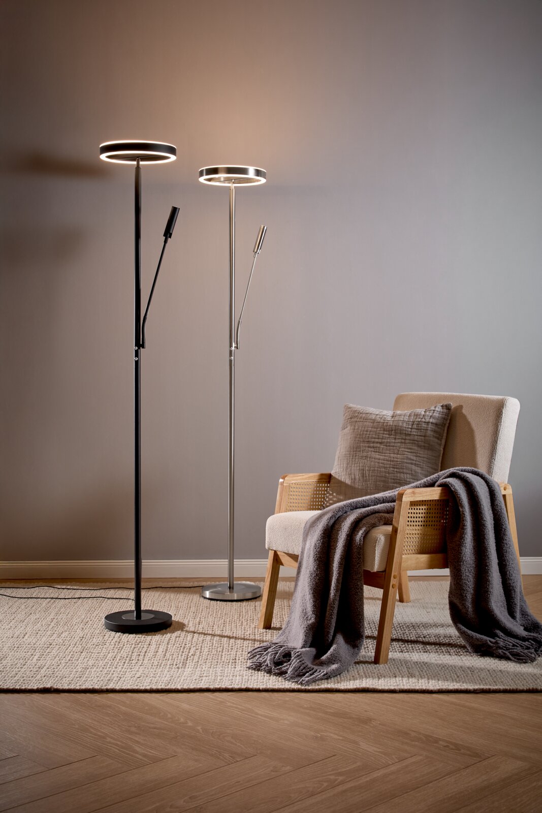 Zwei moderne LED-Deckenfluter-Stehlampen mit ringförmigem Leuchtkopf und zusätzlichem Lesearm (schwarz und silber) neben einem beigen Sessel mit Kissen und grauer Decke, aufgenommen in frontaler Perspektive auf Augenhöhe.