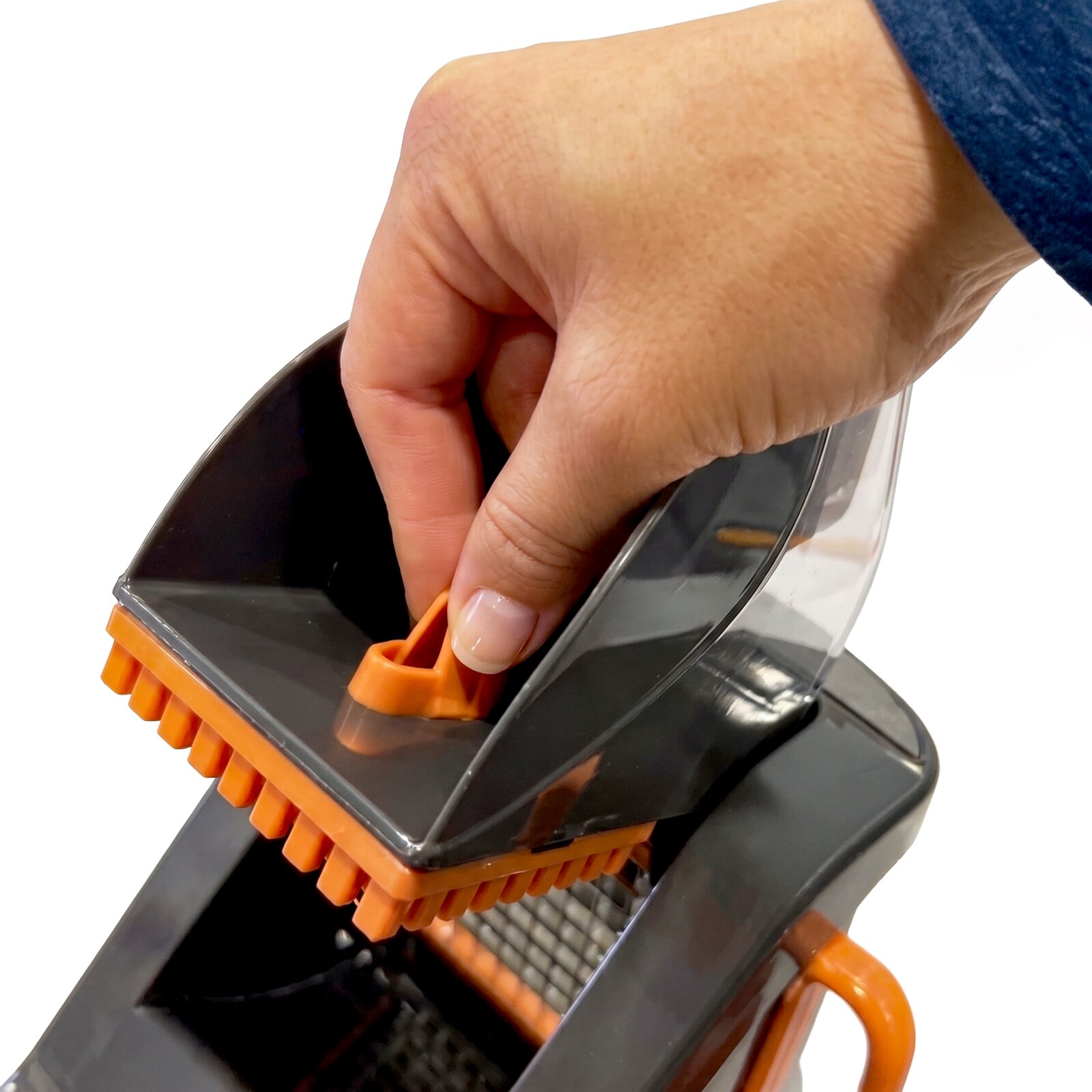 Close-up schuin bovenaanzicht van een hand die de oranje aandrukker van een grijze mandoline/groentesnijder met oranje borstelrand vastpakt op een witte achtergrond
