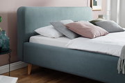Seitliche Ansicht eines modernen Polsterbetts in Blau mit mehreren Kissen und einer weißen Bettdecke, auf einem Holzboden stehend.