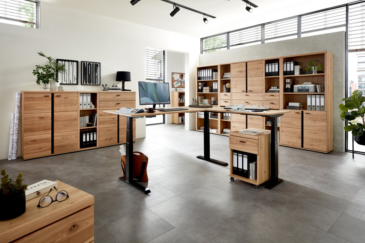 Moderne Büromöbel in einem hellen Raum mit großen Fenstern, bestehend aus Aktenschränken, Regalen und einem höhenverstellbaren Schreibtisch. Perspektive von vorne.