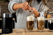 Close-up van twee glazen latte macchiato met melkschuim, die op een houten plank staan, terwijl iemand er chocoladeschilfers overheen strooit. Ook is er een zwarte melkopschuimer in beeld te zien.