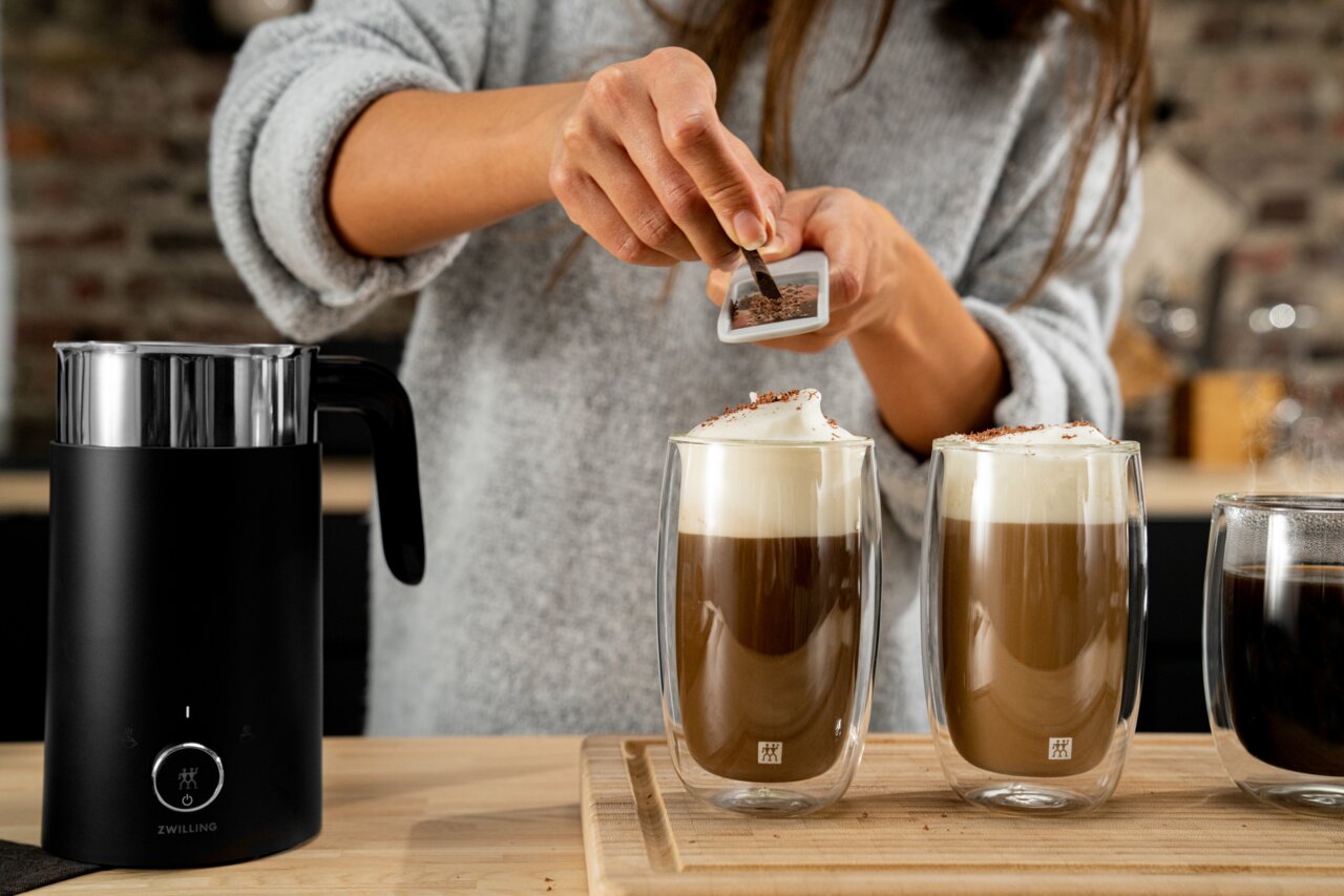 Close-up van twee glazen latte macchiato met melkschuim, die op een houten plank staan, terwijl iemand er chocoladeschilfers overheen strooit. Ook is er een zwarte melkopschuimer in beeld te zien.