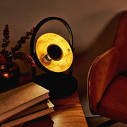 Tischleuchte OLE in einem gemütlichen Wohnzimmer, seitliche Perspektive, warmes Licht, neben einem Sessel und Büchern auf einem Holztisch.