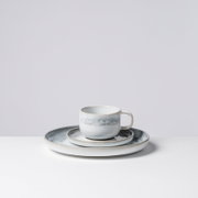 Keramik-Kaffeetasse mit Henkel in Weiß-Blau mit zarter marmorierter Glasur, auf passender Untertasse und großem Teller, freigestellt vor hellem Hintergrund, leicht seitliche Frontansicht von oben