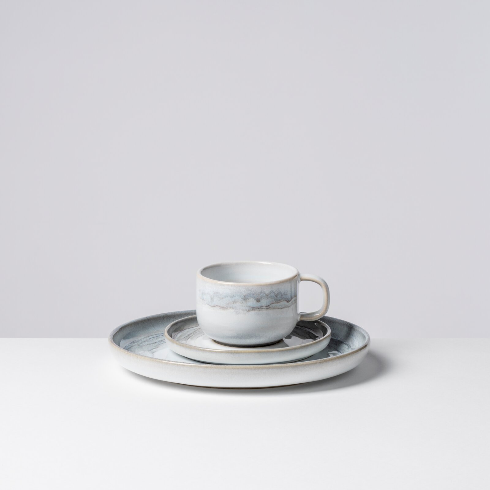 Keramik-Kaffeetasse mit Henkel in Weiß-Blau mit zarter marmorierter Glasur, auf passender Untertasse und großem Teller, freigestellt vor hellem Hintergrund, leicht seitliche Frontansicht von oben
