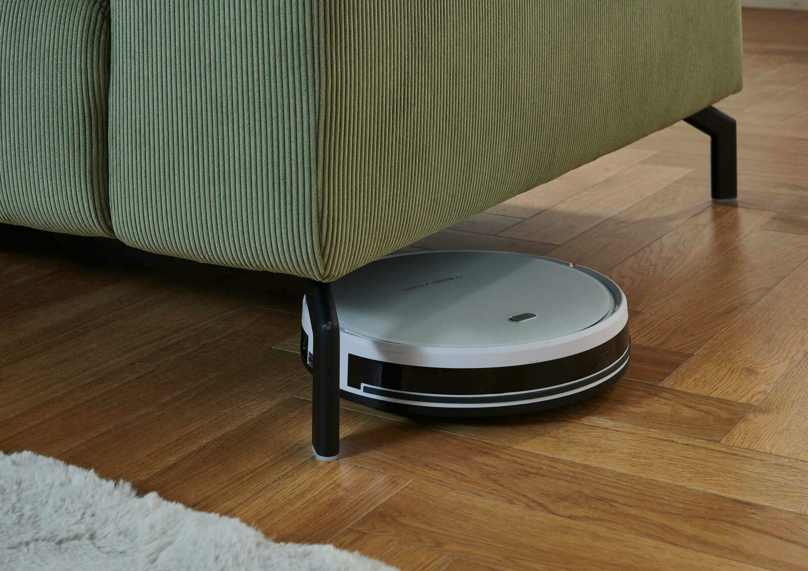 Ein Saugroboter reinigt den Boden unter einer grünen Eckgarnitur aus der Seitenperspektive.