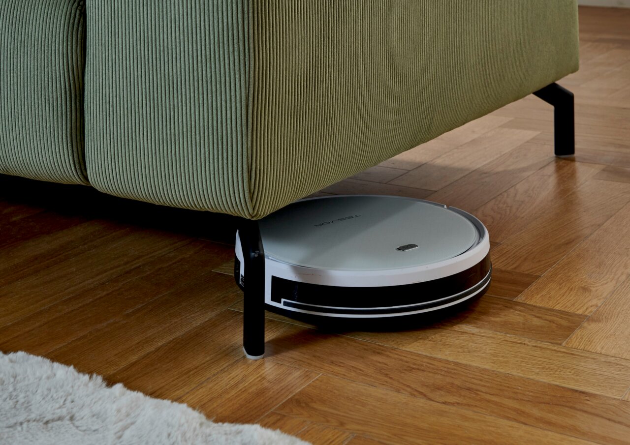 Ein Saugroboter reinigt den Boden unter einer grünen Eckgarnitur aus der Seitenperspektive.
