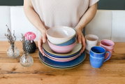 Person hält ein gestapeltes Set aus buntem Keramikgeschirr mit Schalen und Tellern in Blau- und Rosatönen auf einem Holztisch; daneben farbige Becher und kleine Glasvasen mit Blumen, Perspektive: frontale Ansicht auf Tischhöhe.