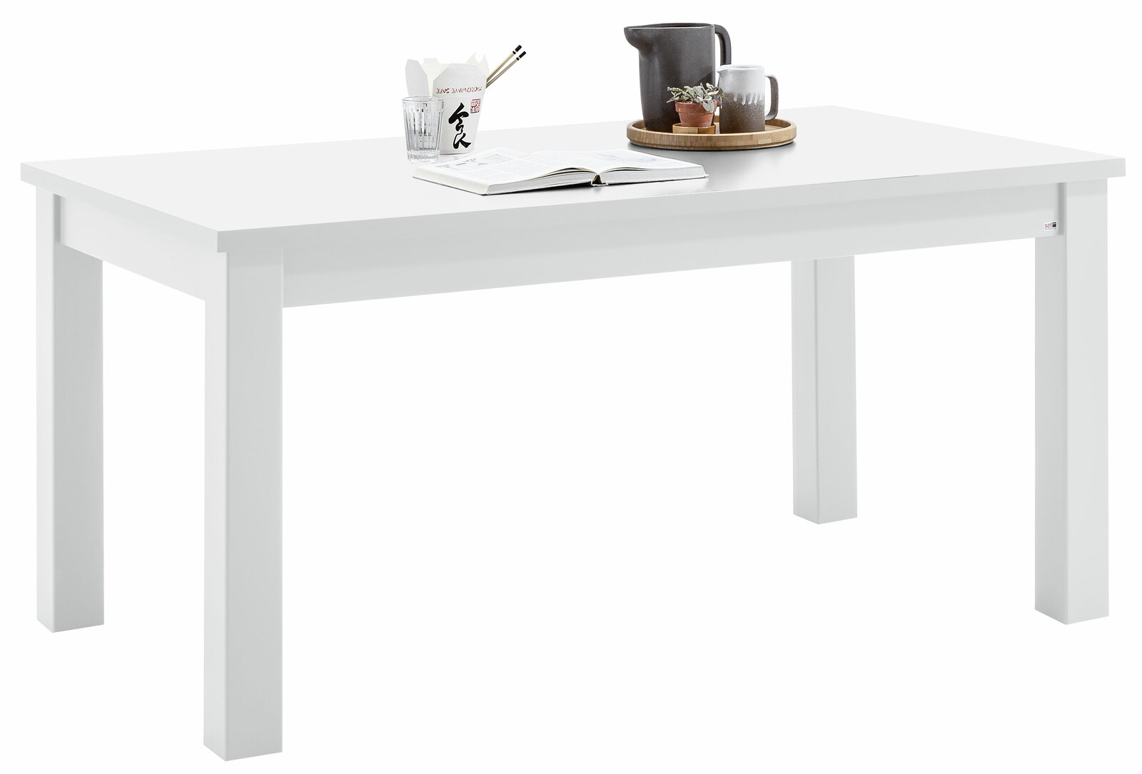 Set one by Musterring Esstisch TACOMA Weißer Esstisch aus der Serie Set one by Musterring, Modell TACOMA, mit Dekorationen wie einem Buch, einer Tasse und einer kleinen Pflanze, aus seitlicher Perspektive fotografiert.