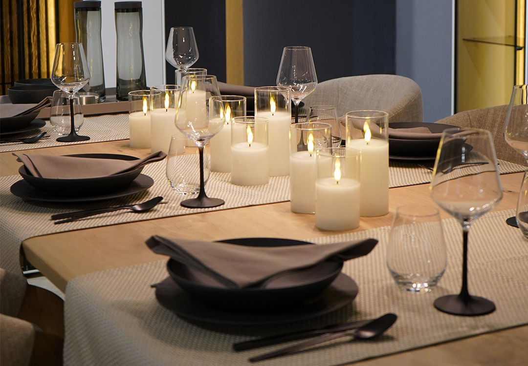 Ein für ein Dinner eingedeckter Esstisch mit Weingläsern, schwarzem Geschirr und vielen Kerzen in der Mitte.