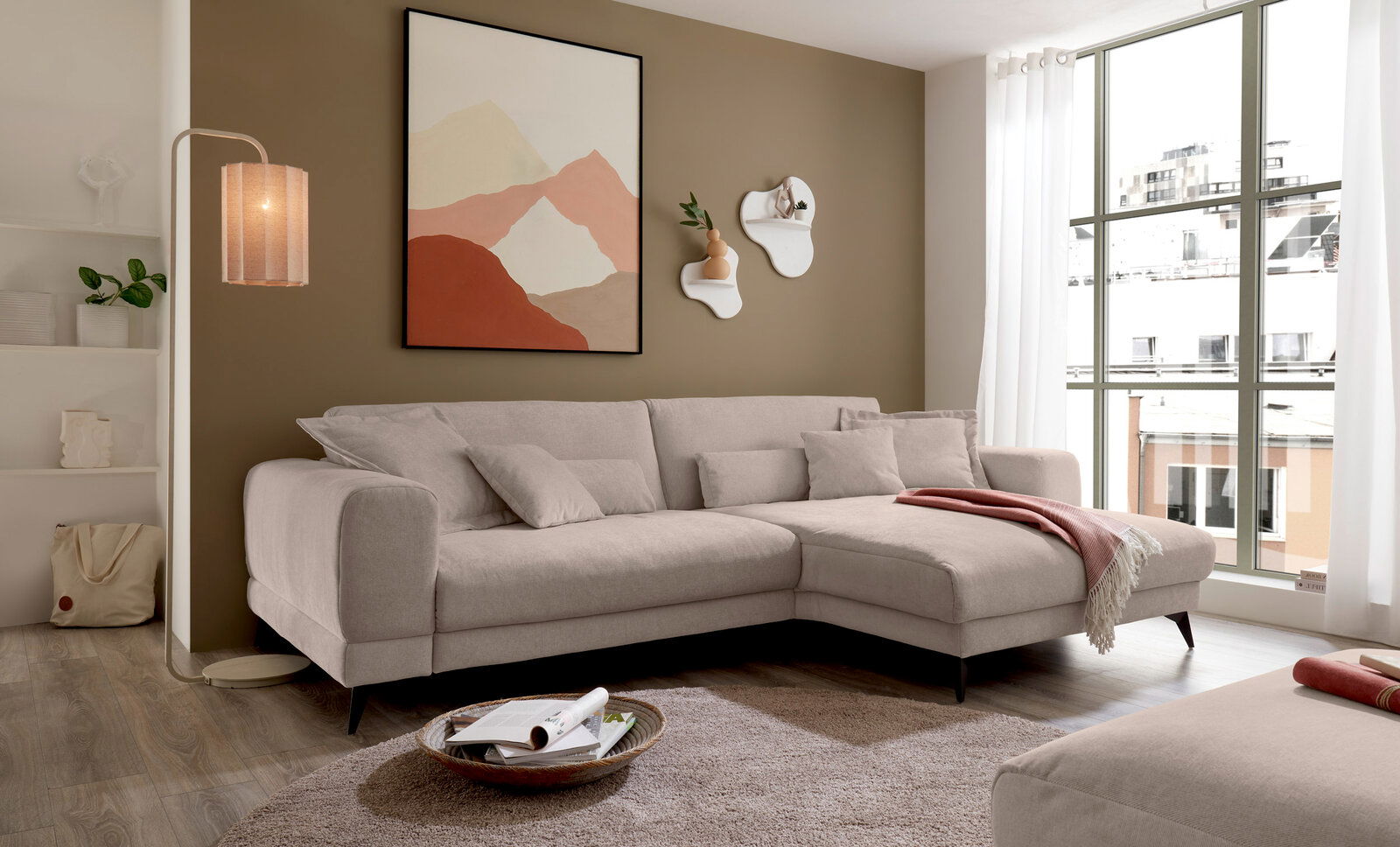 Beige Ecksofa in modernem Wohnzimmer mit großen Fenstern, Wandkunst und dekorativen Regalen, seitliche Perspektive.