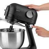 Schwarze Küchenmaschine mit Edelstahl-Rührschüssel, seitliche Ansicht, Hände bedienen die Maschine.