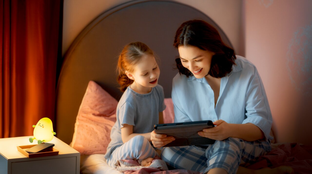 Een accu-nachtlampje in de vorm van een vrolijk, lichtgevend figuurtje staat op een nachtkastje naast een smartphone. Op de achtergrond zitten een vrouw en een kind op een bed en kijken samen naar een tablet. De scène is vanuit een zijdelings perspectief 