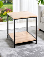 Quadratischer Beistelltisch im Industrial-Design mit heller Holzoptik und schwarzem Metallrahmen sowie Ablageboden, fotografiert in schräger Vorderansicht im Wohnzimmer neben einem Sofa.