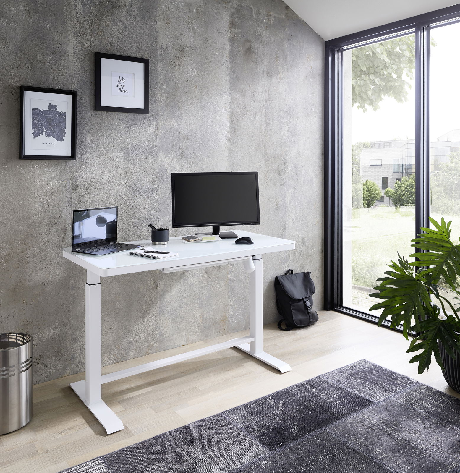 Moderner weißer E-Schreibtisch in einem hellen Raum mit Blick auf einen Garten, ausgestattet mit einem Monitor, Laptop und Schreibtischzubehör, seitliche Perspektive