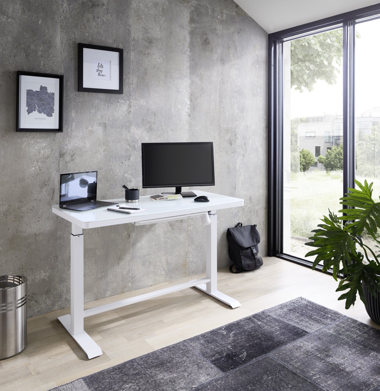 Moderner weißer E-Schreibtisch in einem hellen Raum mit Blick auf einen Garten, ausgestattet mit einem Monitor, Laptop und Schreibtischzubehör, seitliche Perspektive