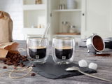 Zwei Espressogläser mit doppelwandigem Design, gefüllt mit Espresso, auf einer Schieferplatte. Im Hintergrund sind Kaffeebohnen, ein Kaffeelöffel und ein Kaffeefilter zu sehen. Perspektive von vorne.