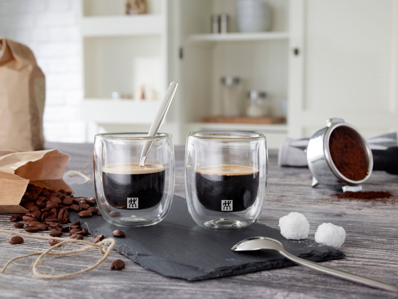 Zwei Espressogläser mit doppelwandigem Design, gefüllt mit Espresso, auf einer Schieferplatte. Im Hintergrund sind Kaffeebohnen, ein Kaffeelöffel und ein Kaffeefilter zu sehen. Perspektive von vorne.