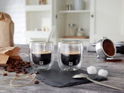 Zwei Espressogläser mit doppelwandigem Design, gefüllt mit Espresso, auf einer Schieferplatte. Im Hintergrund sind Kaffeebohnen, ein Kaffeelöffel und ein Kaffeefilter zu sehen. Perspektive von vorne.
