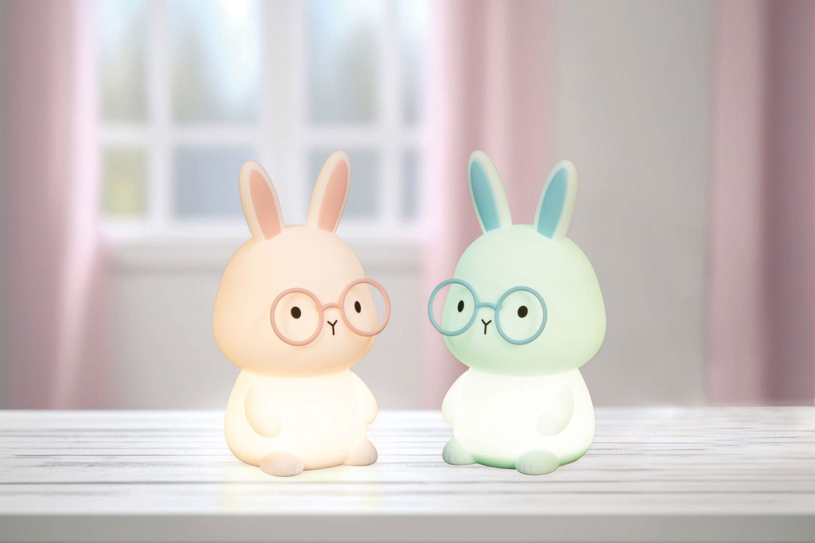 Zwei leuchtende Nachtlichter in Form von Hasen mit Brillen, auf einem Tisch vor einem Fenster, frontal betrachtet.